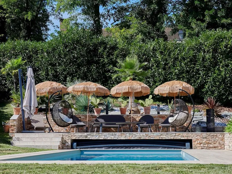 Jardin paysagé autour d'une piscine : parasols en paille, fauteuils suspendus, muret en pierre sèche et massifs plantés — l'eau et le végétal composés ensemble