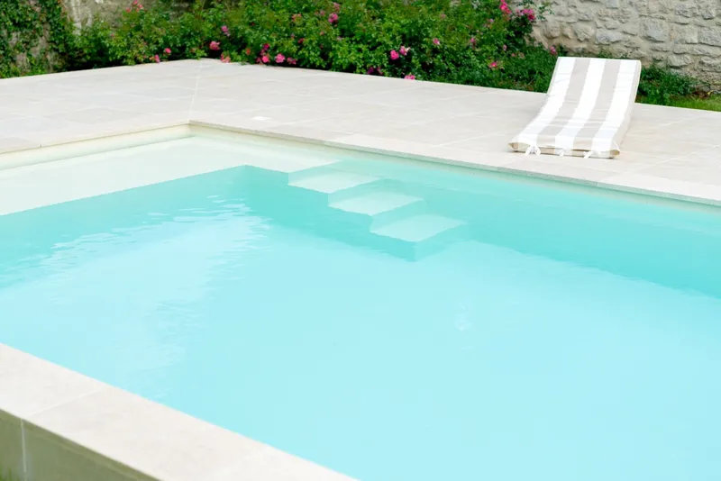 Gros plan sur l'angle d'un bassin à l'eau turquoise, margelles et plage en pierre claire, escalier immergé, transat à rayures, rosiers grimpants et mur de pierre ancienne en fond — réalisation Aqua System, ouest parisien