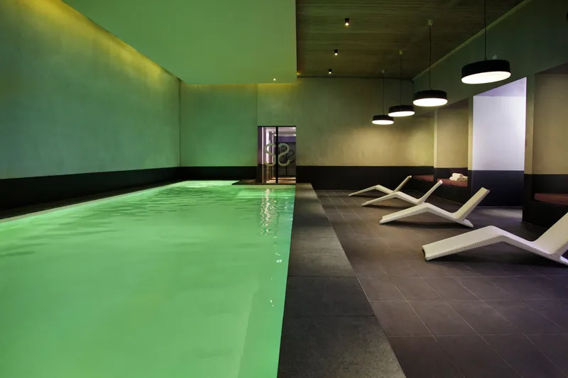 Bassin intérieur en eau, parois enduites baignées d'une lumière d'ambiance verte, sol et plinthe sombres, suspensions noires au-dessus d'une rangée de transats blancs, porte vitrée au fond — réalisation Aqua System, ouest parisien