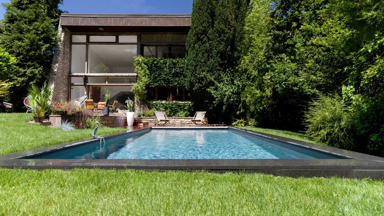 Jardin paysagé avec bassin intégré aux margelles sombres, pelouse soignée et maison à bardage bois habillée de plantes grimpantes, cyprès et massifs — réalisation Les Terres Essentielles dans l'ouest parisien