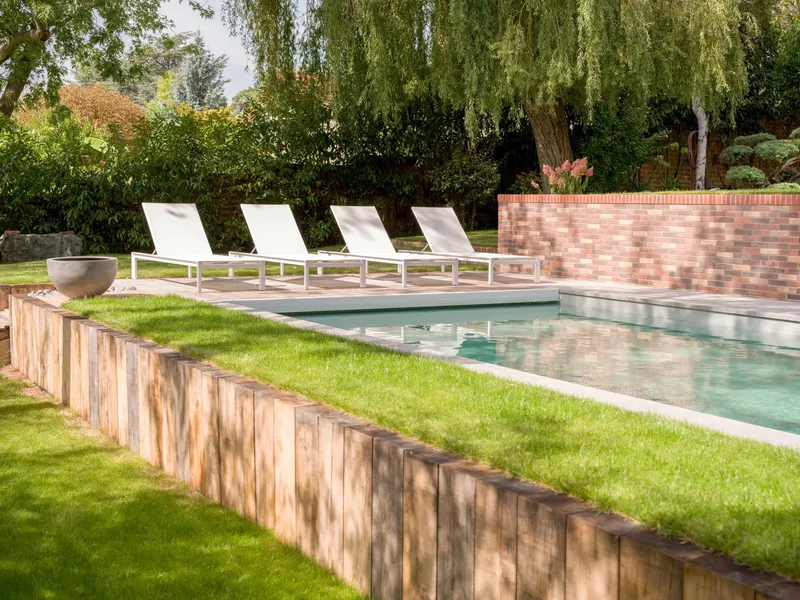 Abords d'une piscine dans un jardin entretenu : pelouse tondue ras, massifs taillés, soutènement en traverses de bois et transats alignés le long du bassin, ouest parisien