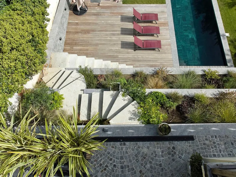 Vue plongeante sur un jardin en terrasses étagées : terrasse en bois exotique et transats au bord d'un couloir de nage à l'eau sombre en haut, volées d'escaliers en pierre claire et massifs de graminées au centre, sol pavé et plantes graphiques en bas — création paysagère Les Terres Essentielles, ouest parisien