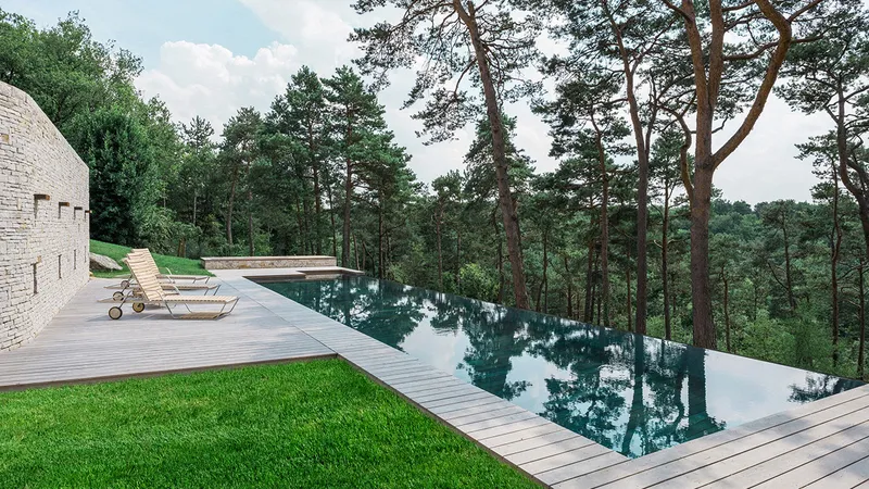 Piscine à débordement bordée d'une terrasse en bois et d'une pelouse, plan d'eau ouvert sur une forêt de pins, mur en pierre, transats au bord — réalisation Aqua System, ouest parisien