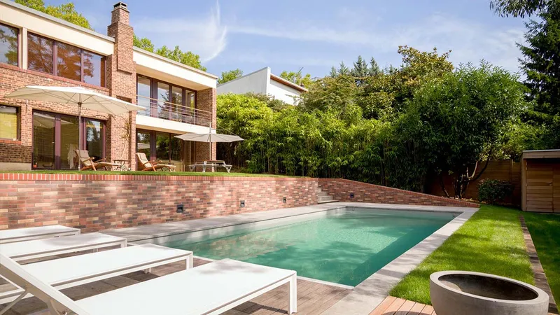 Bassin à l'eau verte bordé de pierre, muret en brique, terrasse haute avec parasols, maison contemporaine en brique et bois, haie dense — réalisation Aqua System, ouest parisien