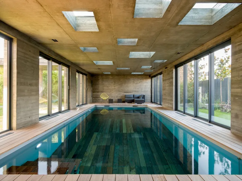 Piscine intérieure en béton brut éclairée par des puits de lumière, plage en bois clair et baies vitrées sur le jardin — ouvrage Aqua System