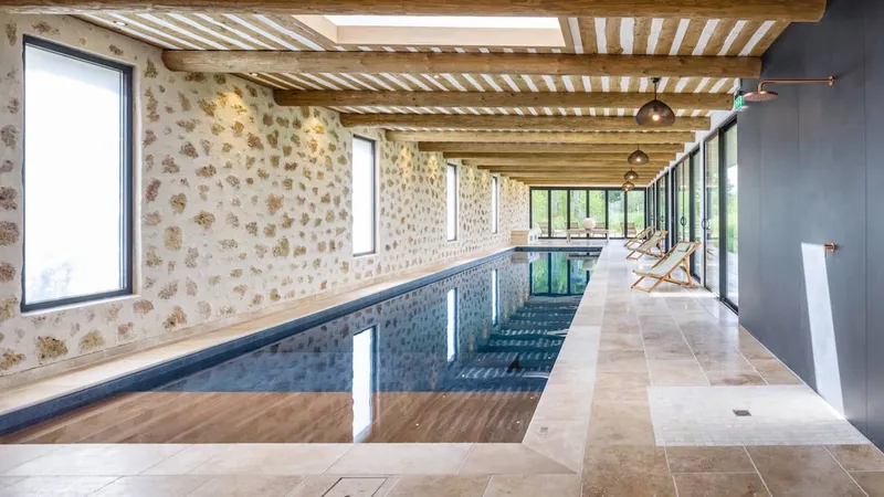 Long couloir de nage intérieur, murs en pierre apparente, plafond à poutres de bois, dallage en travertin, baies vitrées sur le jardin, transats — réalisation Aqua System, ouest parisien