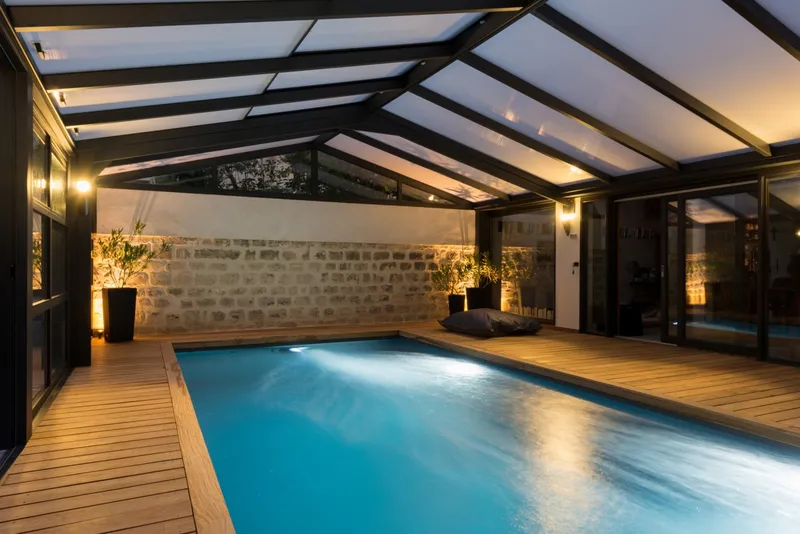 Piscine couverte sous une véranda à charpente métallique noire et toit vitré incliné, mur en pierre ancienne mis en lumière, terrasse de bois ceinturant le bassin, eau bleutée, éclairage chaud de soirée — réalisation Aqua System, ouest parisien