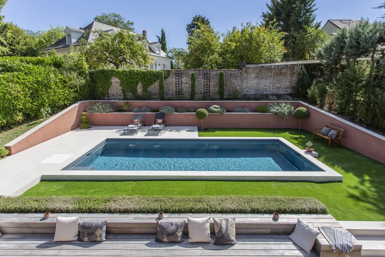 Piscine au revêtement gris-bleu en plein soleil dans un jardin clos de murs anciens en brique, terrasses en pierre claire, transats et massifs taillés — réalisation Aqua System, ouest parisien