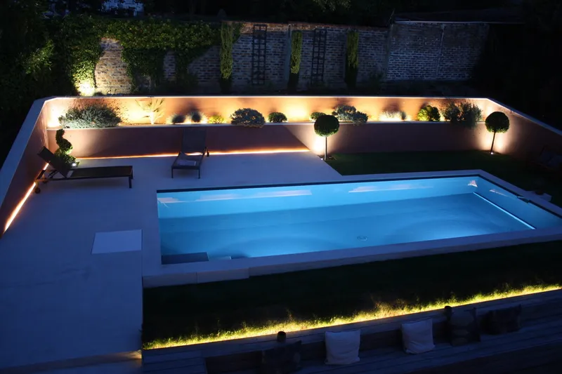 Vue plongeante nocturne sur un bassin rectangulaire éclairé en bleu, terrasse minérale claire, murets et bandes de pelouse soulignés d'un éclairage indirect, mur de pierre ancienne en fond, banquette à coussins au premier plan — projet complet eau et jardin Aqua System, ouest parisien