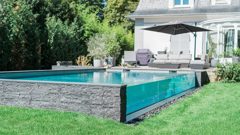 Piscine surélevée à paroi de verre transparente, parement en pierre grise empilée, pelouse, salon de jardin et parasol devant une maison — réalisation Aqua System, ouest parisien