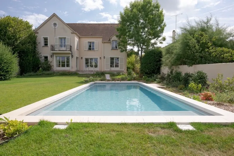 Bassin rectangulaire à margelles de pierre claire au milieu d'une grande pelouse, devant une demeure beige à lucarnes et volets, massifs et arbres de part et d'autre — projet complet eau et jardin Aqua System, ouest parisien