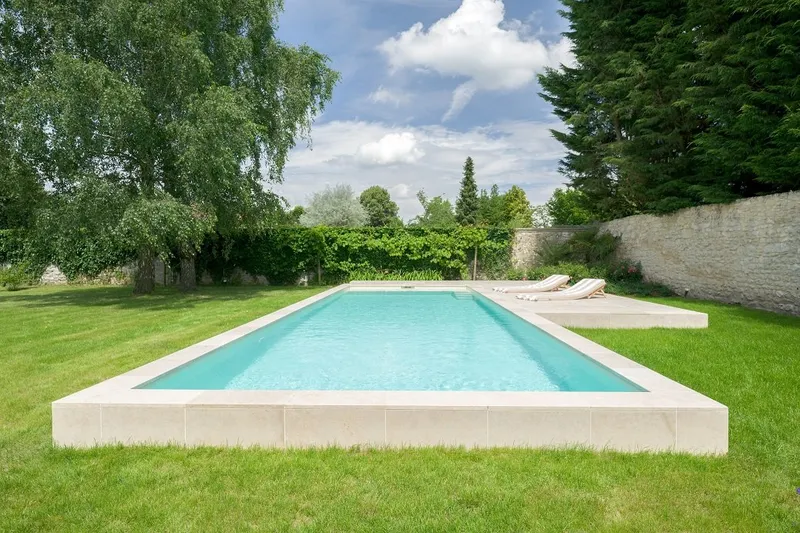 Piscine bordée de margelles en pierre claire le long d'un mur ancien restauré, abords minéraux soignés — réalisation Aqua System, ouest parisien