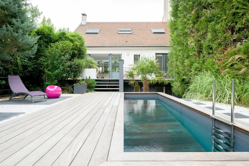 Bassin sombre encastré dans une large terrasse de bois, devant une maison contemporaine à lucarnes, assises colorées et bac planté au premier plan, hautes graminées et mur végétal de part et d'autre — projet complet eau et jardin Aqua System, ouest parisien