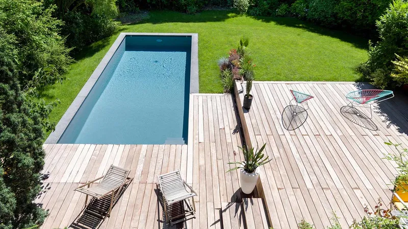 Vue plongeante sur un bassin rectangulaire bordé d'une vaste terrasse en bois, pelouse, transats et fauteuils design, végétation périphérique — réalisation Aqua System, ouest parisien