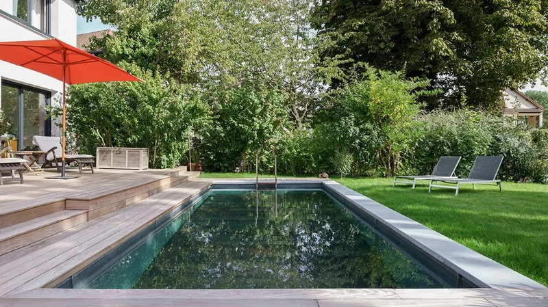 Bassin rectangulaire en béton dans un jardin clos structuré, pelouse, banquette de terrasse en bois avec coussins, demeure ancienne en arrière-plan — projet complet eau et jardin Aqua System, ouest parisien