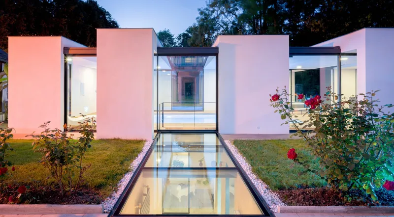 Pavillon de piscine aux volumes blancs et baies vitrées, passerelle de verre posée au sol au-dessus de l'eau, massifs de rosiers rouges et bandes de pelouse de part et d'autre, lumière de fin de journée — projet complet eau et jardin Aqua System, ouest parisien